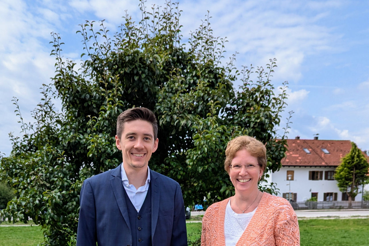 Ein Mann und eine Frau in Geschäftskleidung posieren im Freien vor der Kulisse eines Wohngebiets.