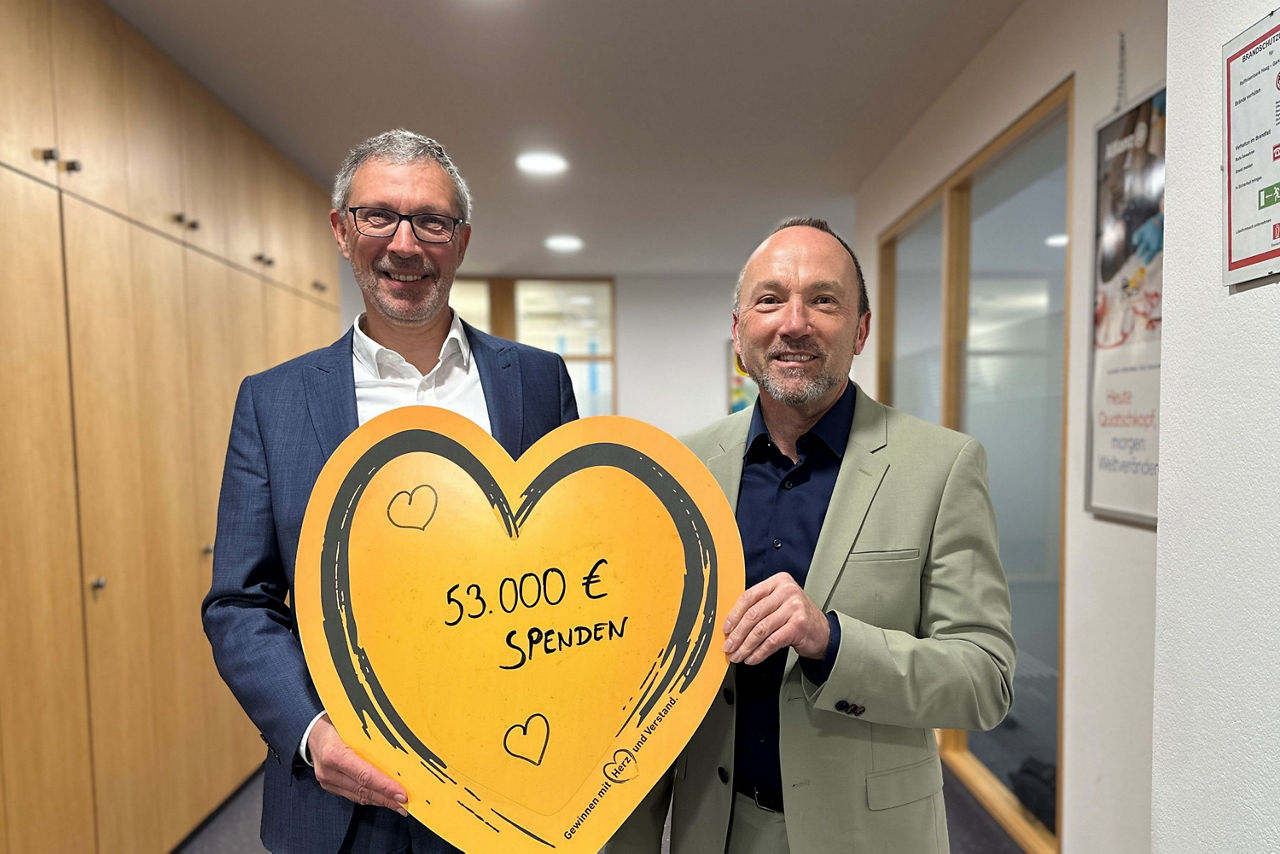 Zwei Männer in Anzügen stehen in einem Büro-Flur und halten ein gelbes Pappherz mit der Aufschrift "53.000 € Spenden"
