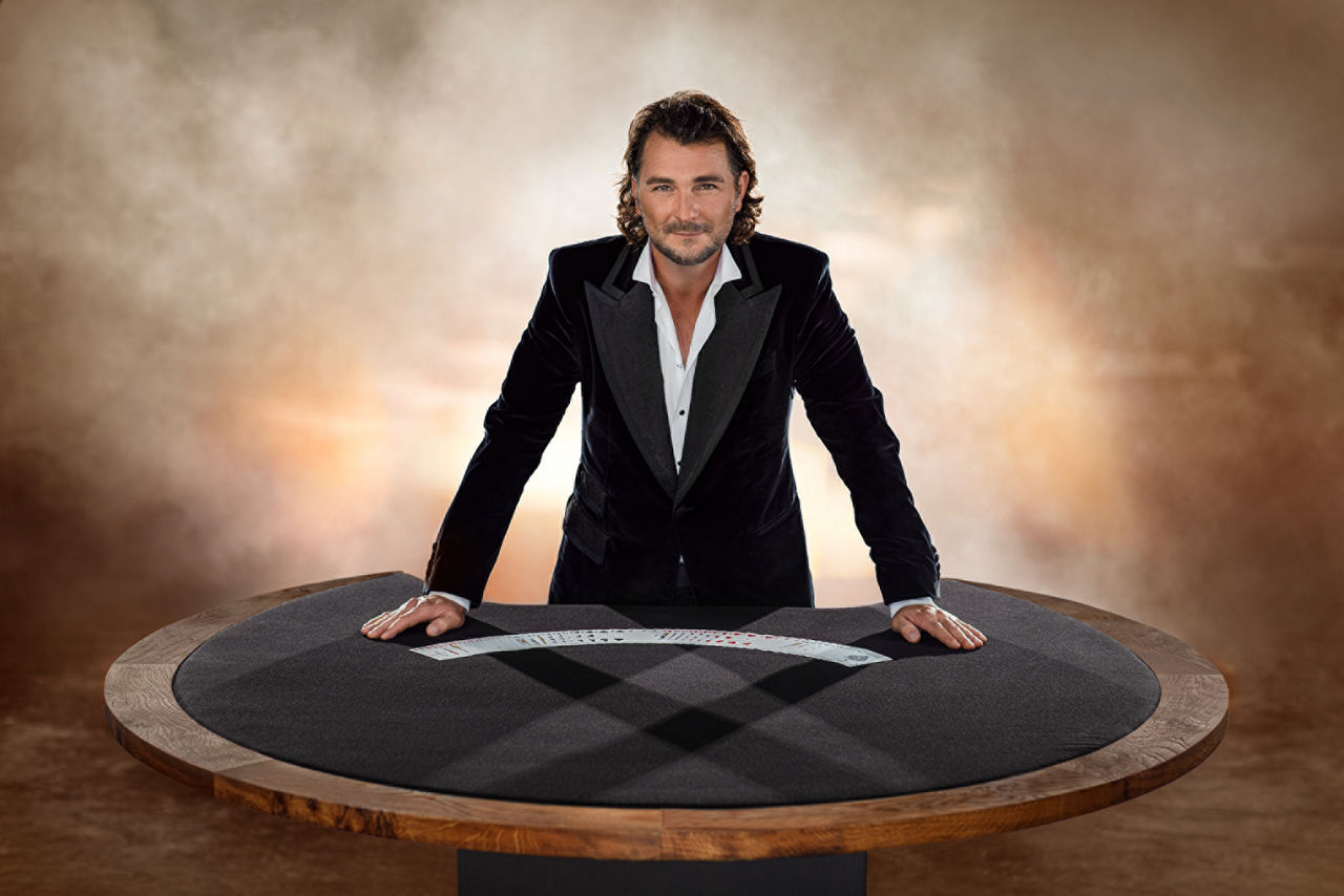 Der bekannte Magier Thorsten Strotmann steht an einem runden Zaubertisch und hat Spielkarten in einem Fächer vor sich liegen.