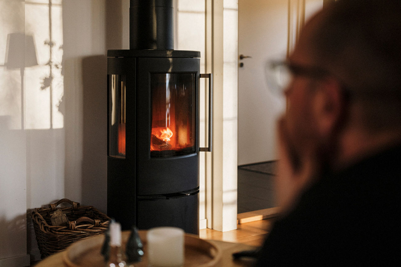 Ein Mann in wohnlicher Atmosphäre beobachtet ein Holzfeuer in einem Kaminofen.