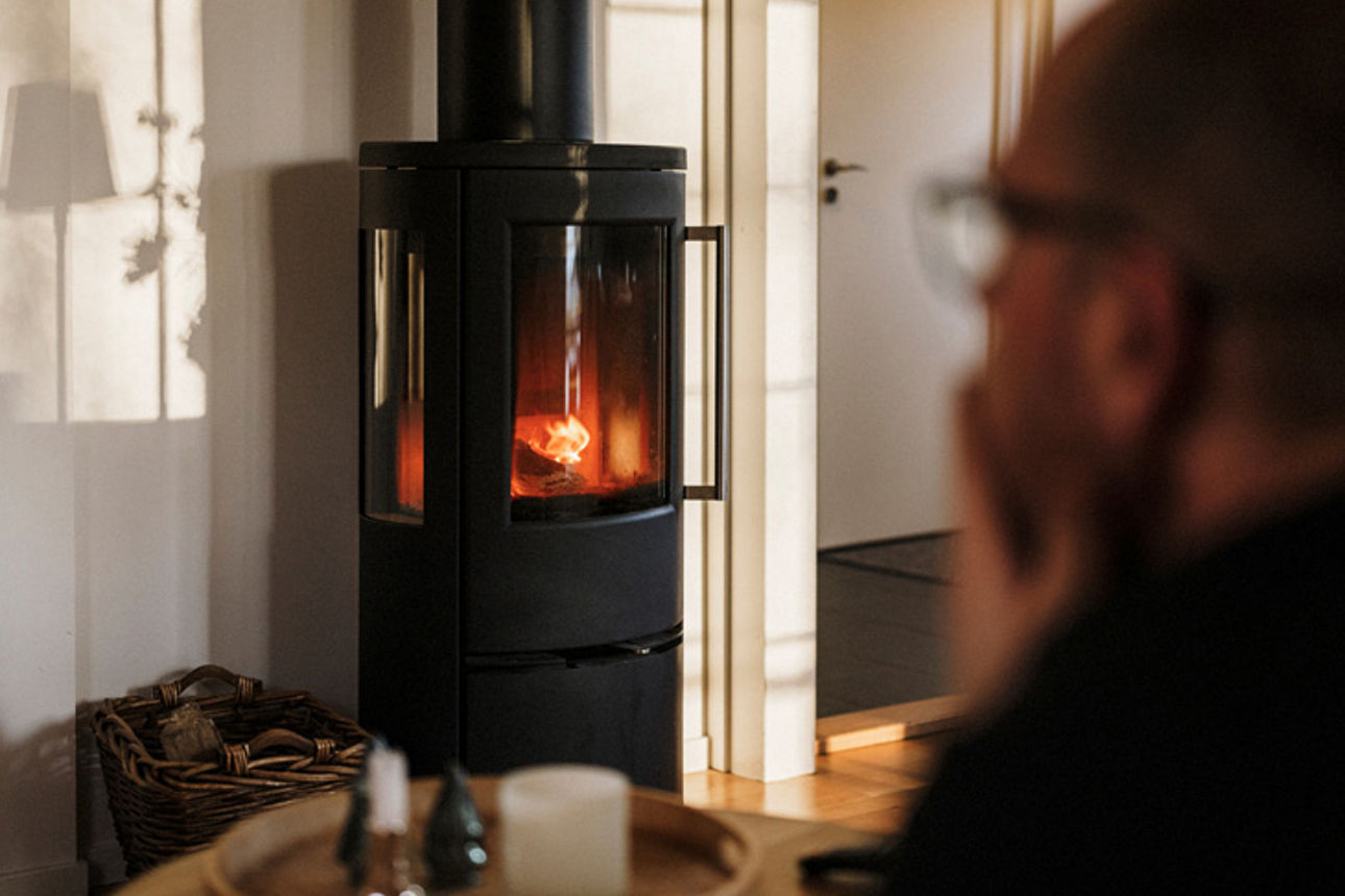 Ein Mann in wohnlicher Atmosphäre beobachtet ein Holzfeuer in einem Kaminofen.