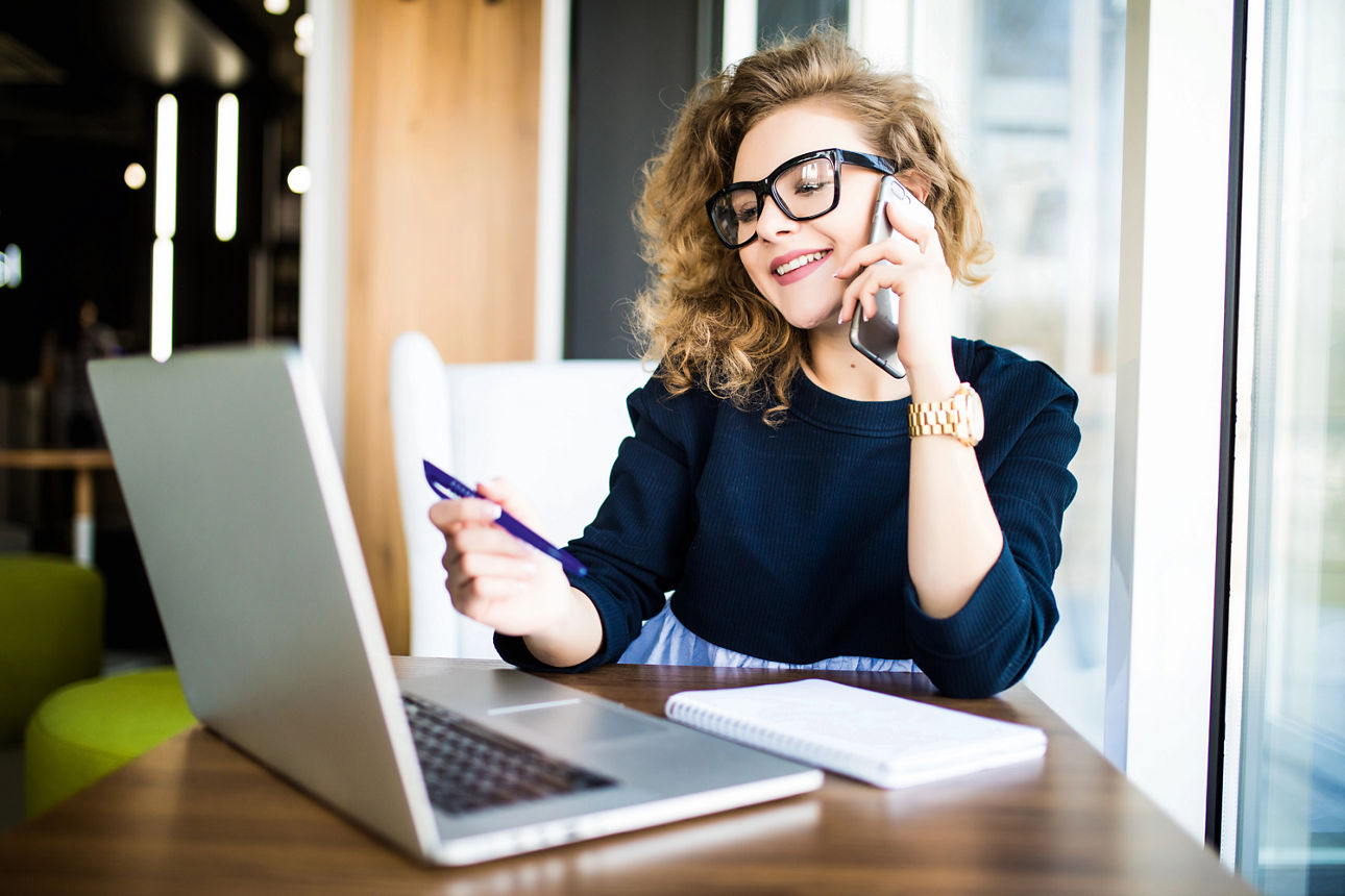 Frau mit Handy am Ohr sitzt an einem Tisch und schaut auf ihren aufgeklappten Laptop