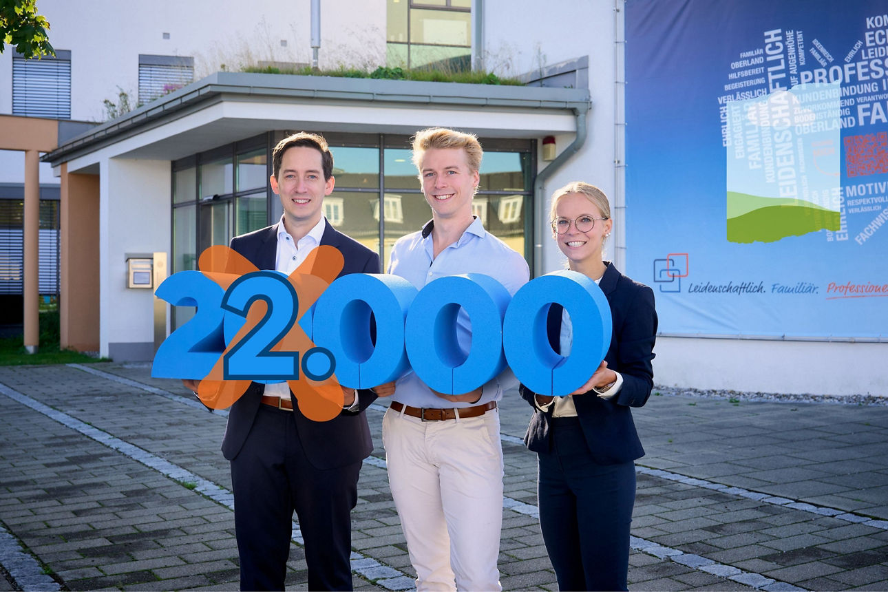Drei Personen posieren mit großen blauen Zahlen, die 20.000 bilden. Im Hintergrund ist das Raiffeisenbank im Oberland Gebäude zu sehen.