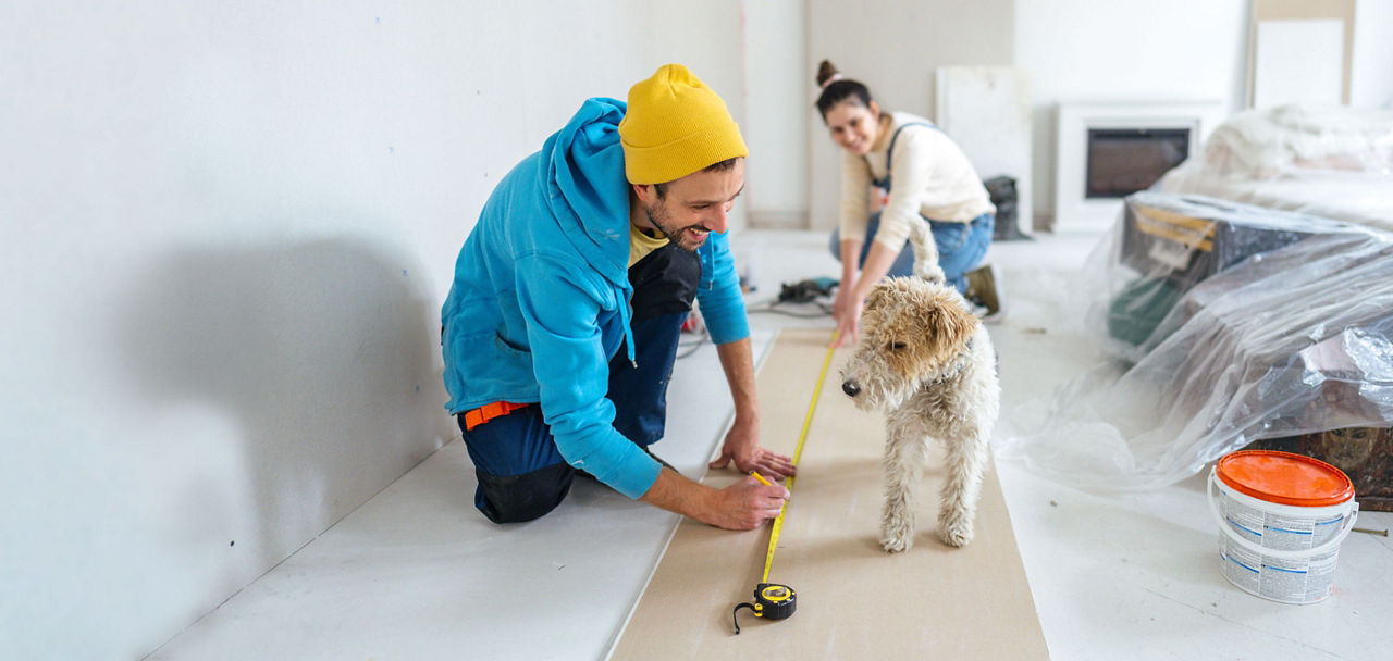 Ein Mann und eine Frau halten ein Maßband auf dem Boden und ein Hund schaut dabei zu