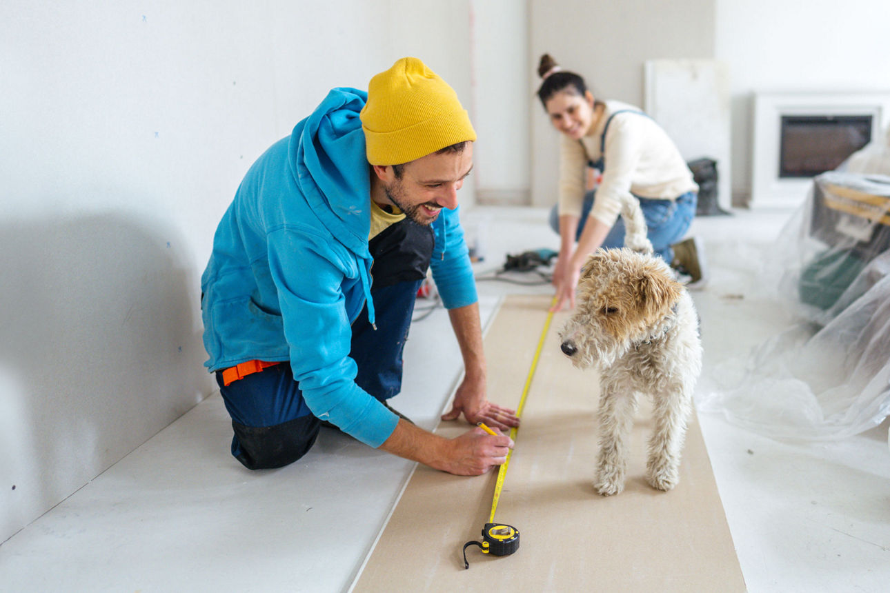 Ein Mann und eine Frau halten ein Maßband auf dem Boden und ein Hund schaut dabei zu