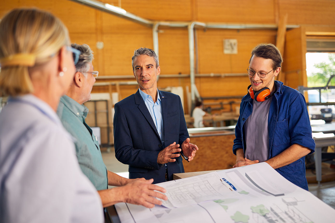 : Vier Personen stehen um einen Tisch mit Bauplänen in einer Werkhalle und diskutieren.