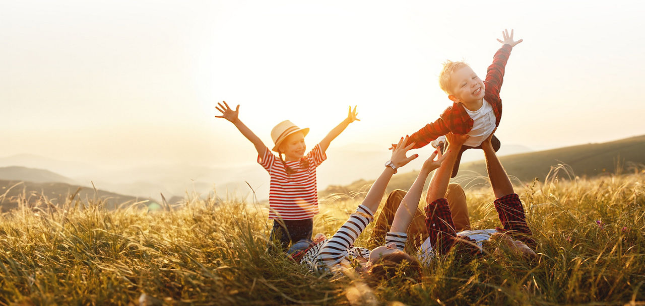 Zwei Erwachsene liegen auf einer Wiese und heben zwei Kinder in die Luft. Die Kinder strecken die Arme zum Fliegen aus.