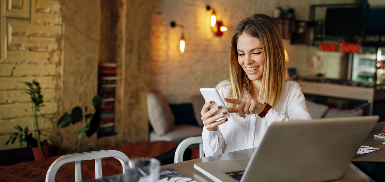 Frau sitzt an einem Tisch mit einem Laptop darauf und hält ein Smartphone in der Hand