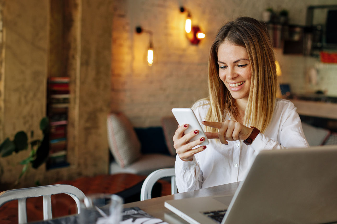 Frau sitzt an einem Tisch mit einem Laptop darauf und hält ein Smartphone in der Hand