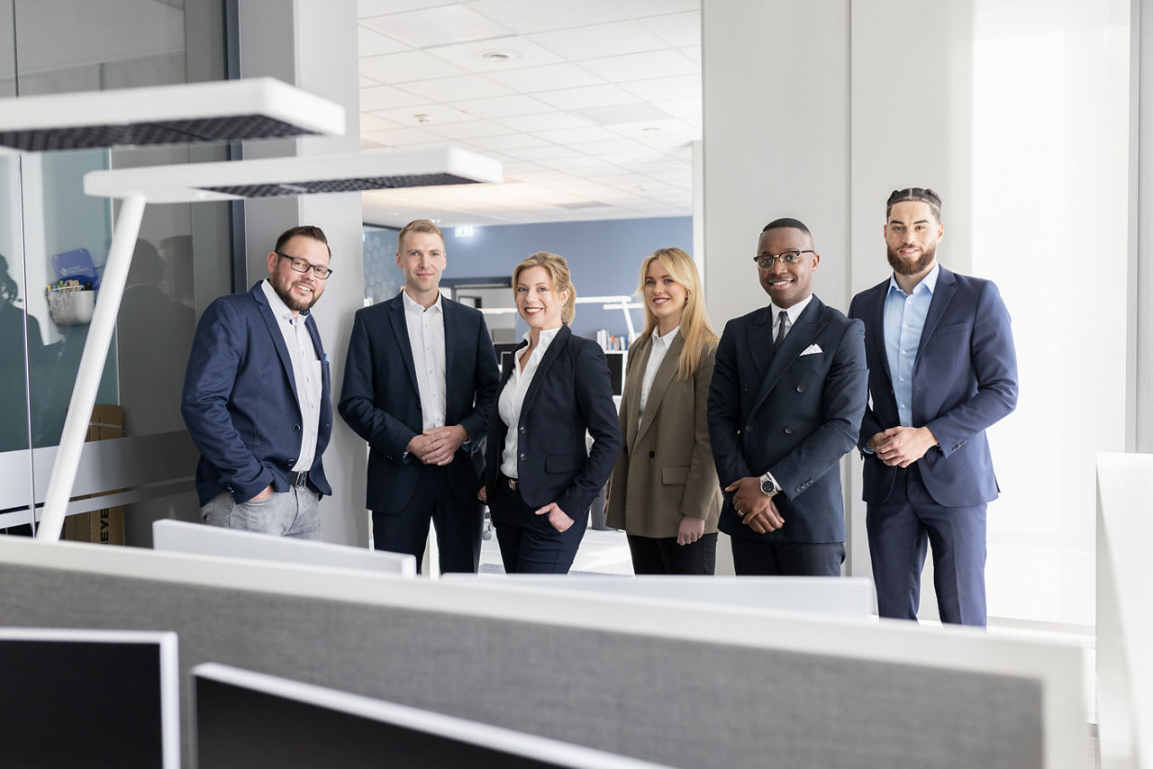 Sechs Mitarbeiterinnen und Mitarbeiter stehen lächelnd im Büro n