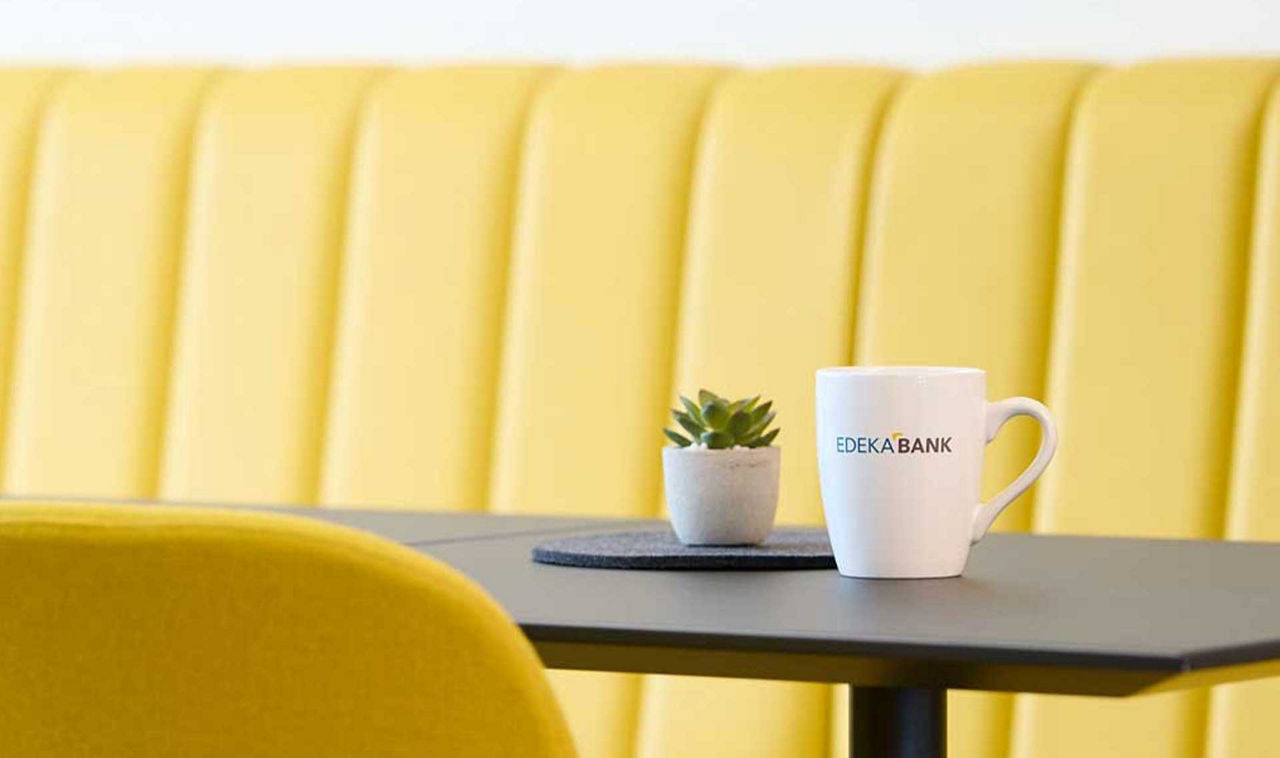 Wir als Arbeitgeber weiße Edekabank Tasse mit Logo vor gelber Sitzecke