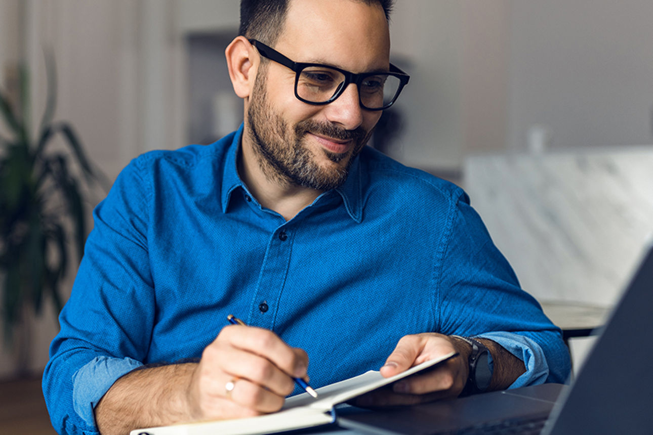 Ein Mann mit einer Brille sitzt vor seinem Laptop und macht sich Notizen auf einem Notizblock