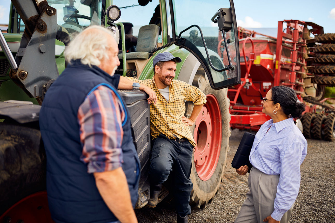 Zwei Landwirte lehnen auf einem Feld an einem Traktor und unterhalten sich mit ihrer Bank-Beraterin