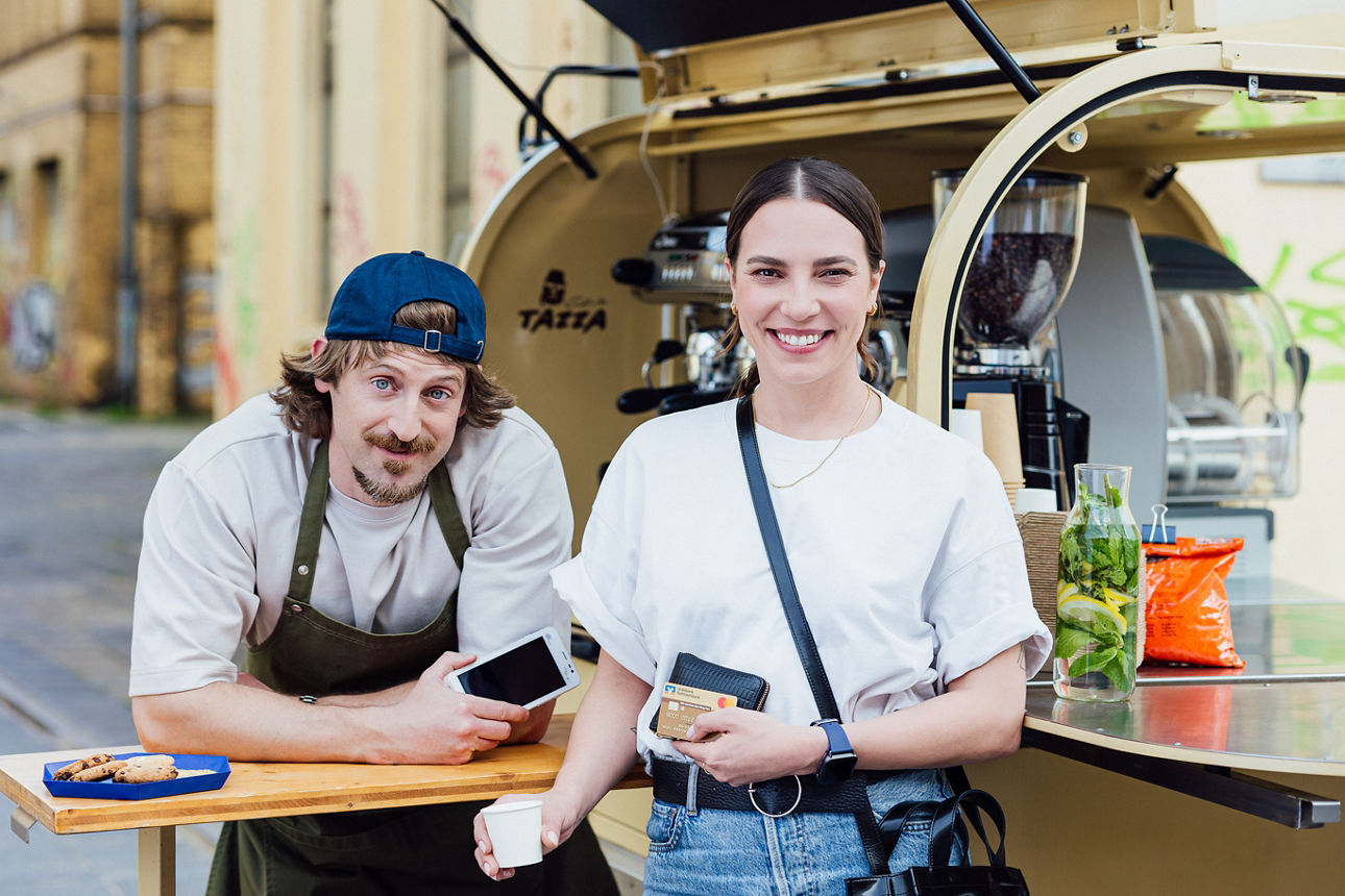 Junge Frau mit Kaffee und Bankkarte steht lächelnd vor einem mobilen Kaffeestand. Daneben ein Barista mit Smartphone.