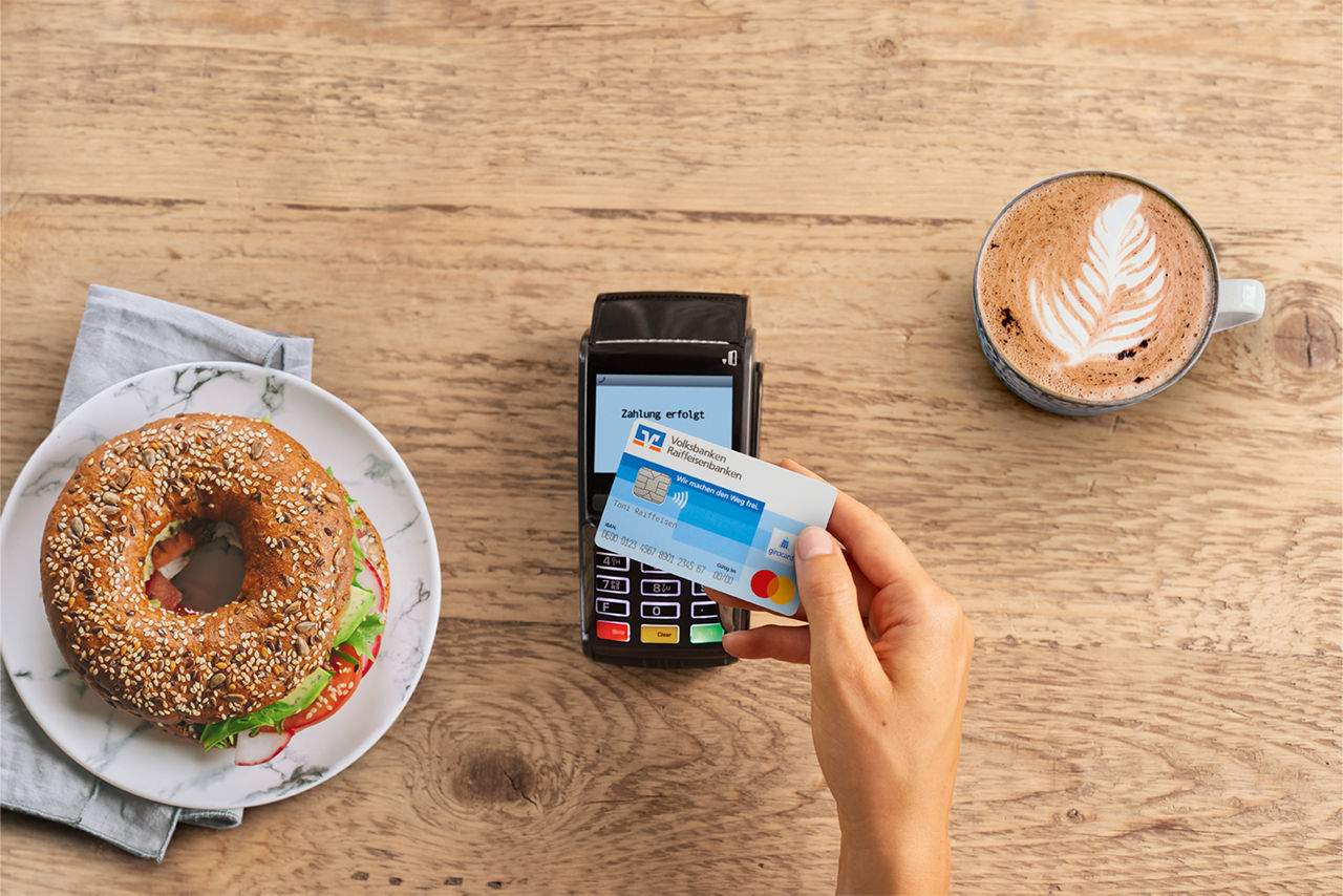 Kontaktloses zahlen in einem Kaffee, Kartenlesegerät und Volksbank Karte neben Bagel und Kaffee