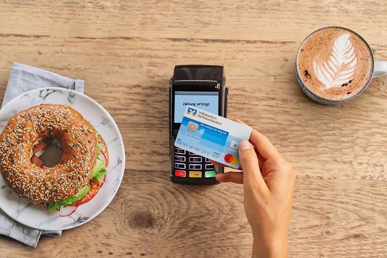 Kontaktloses zahlen in einem Kaffee, Kartenlesegerät und Volksbank Karte neben Bagel und Kaffee