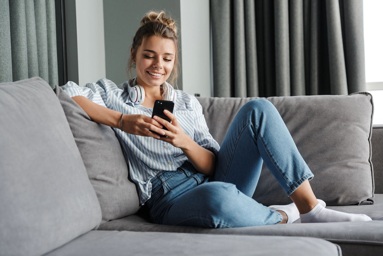Frau mit Handy in beiden Händen sitzt auf einem Sofa und schaut lächelnd auf ihr Handy