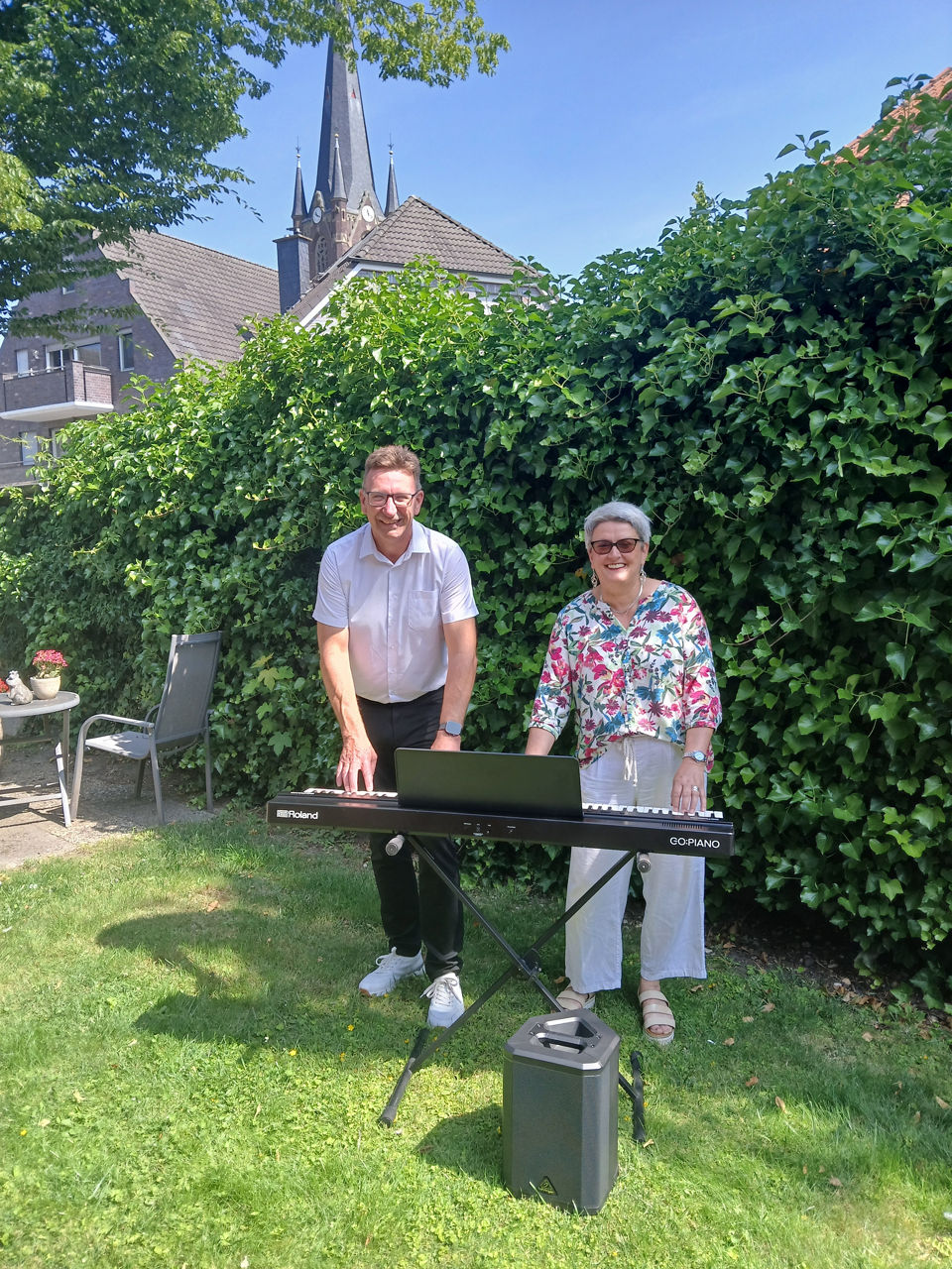 Ein Mitarbeiter der Volksbank und eine Frau vor einer Hecke mit einem e-Piano und einer Box 