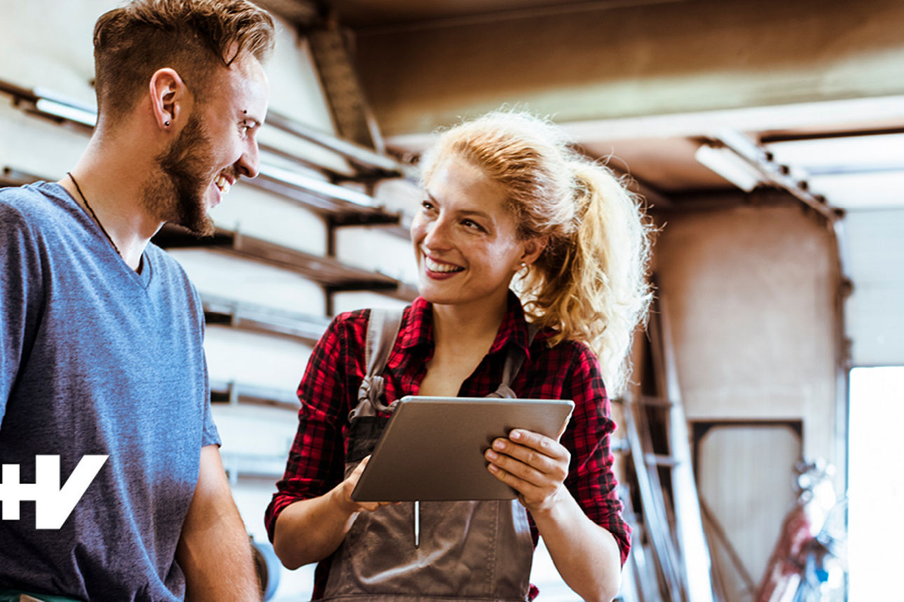 Lachender Mann und lachende Frau in einer Werkstatt, Frau hält ein Tablet in der Hand