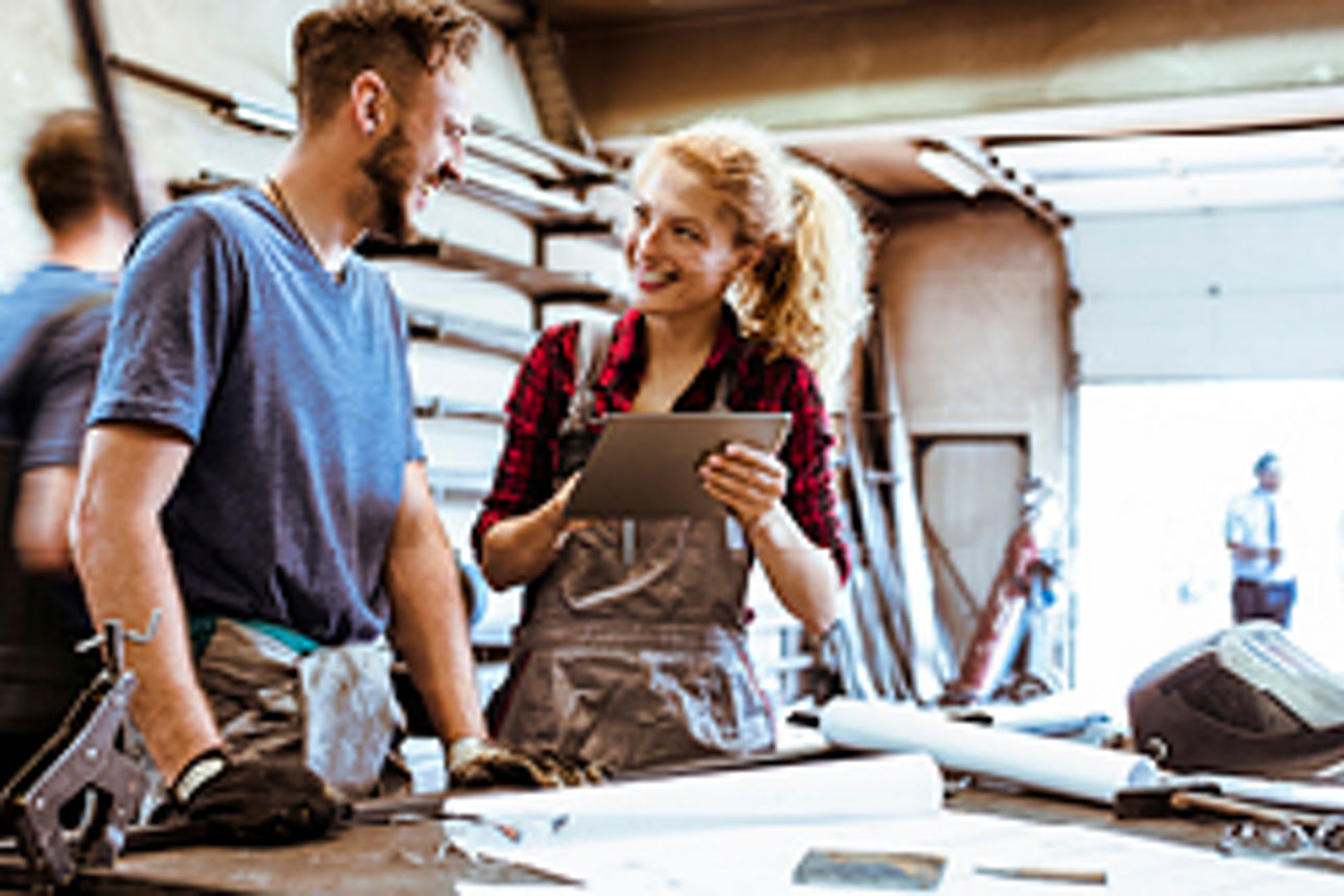 Lachender Mann und lachende Frau in einer Werkstatt, Frau hält ein Tablet in der Hand