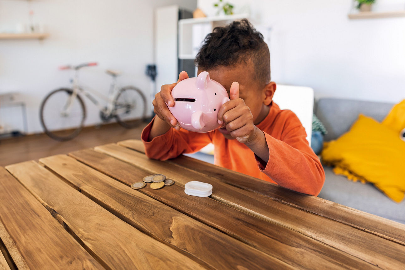 Junge mit orangenem Pullover sitzt am Tisch und leert sein rosanes Sparschwein