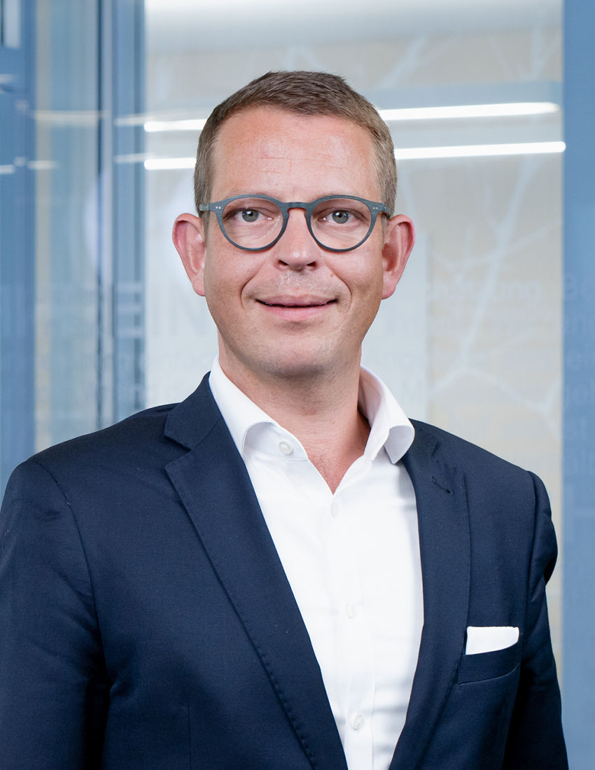 Foto unseres Vermögensberaters Christoph Arend vor einer Büroglaswand in einem dunkelblauen Sakko