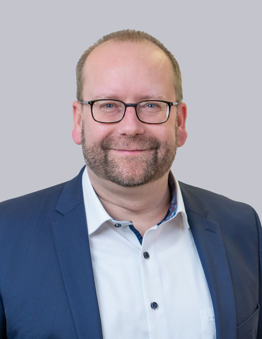 Foto unseres Finanzberaters Patrick Schönig-Roth vor einem grauen  Hintergrund in einem blauen Sakko
