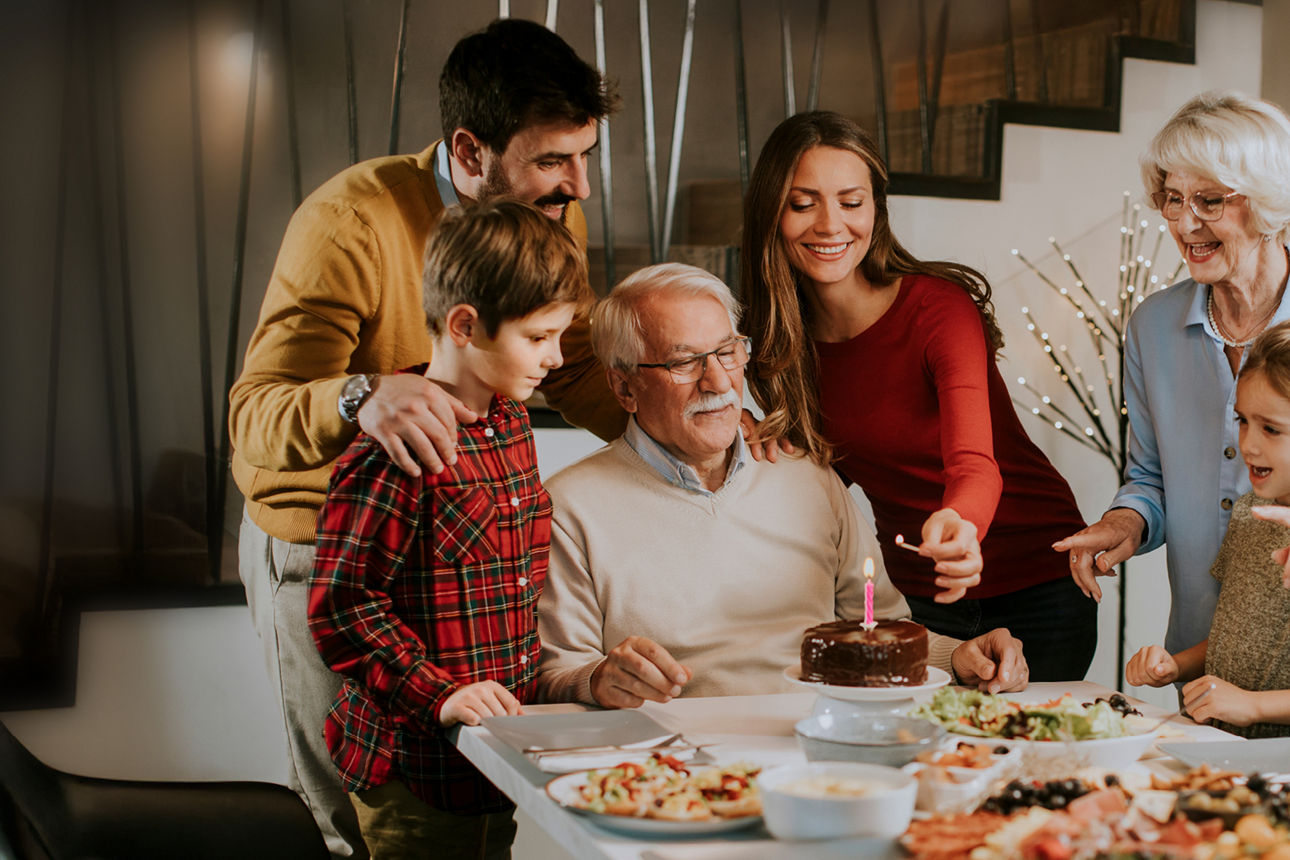 .Eine Familie feiert Geburtstag mit einer Torte an einem Tisch. Die Frau zündet die Kerze an.