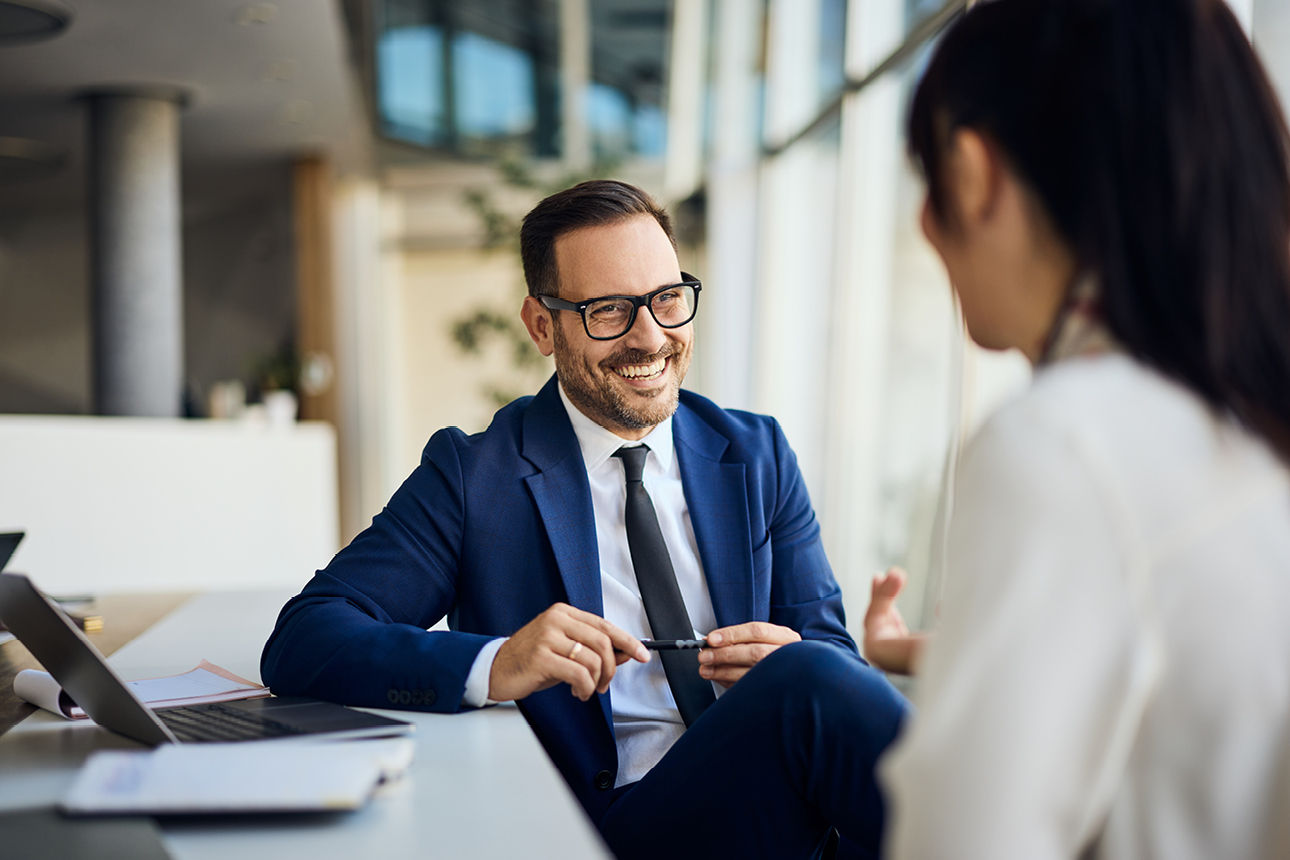 Ein Mann mit Brille und dunkelblauem Anzug sitzt an einem Tisch und hält ein Beratungsgespräch.