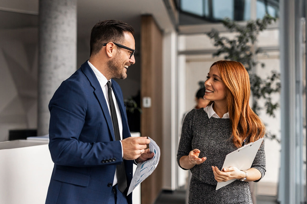 Zwei Kollegen unterhalten sich. Der Mann trägt eine Brille und einen dunkelblauen Anzug. Die Dame trägt einen braunen Pullove