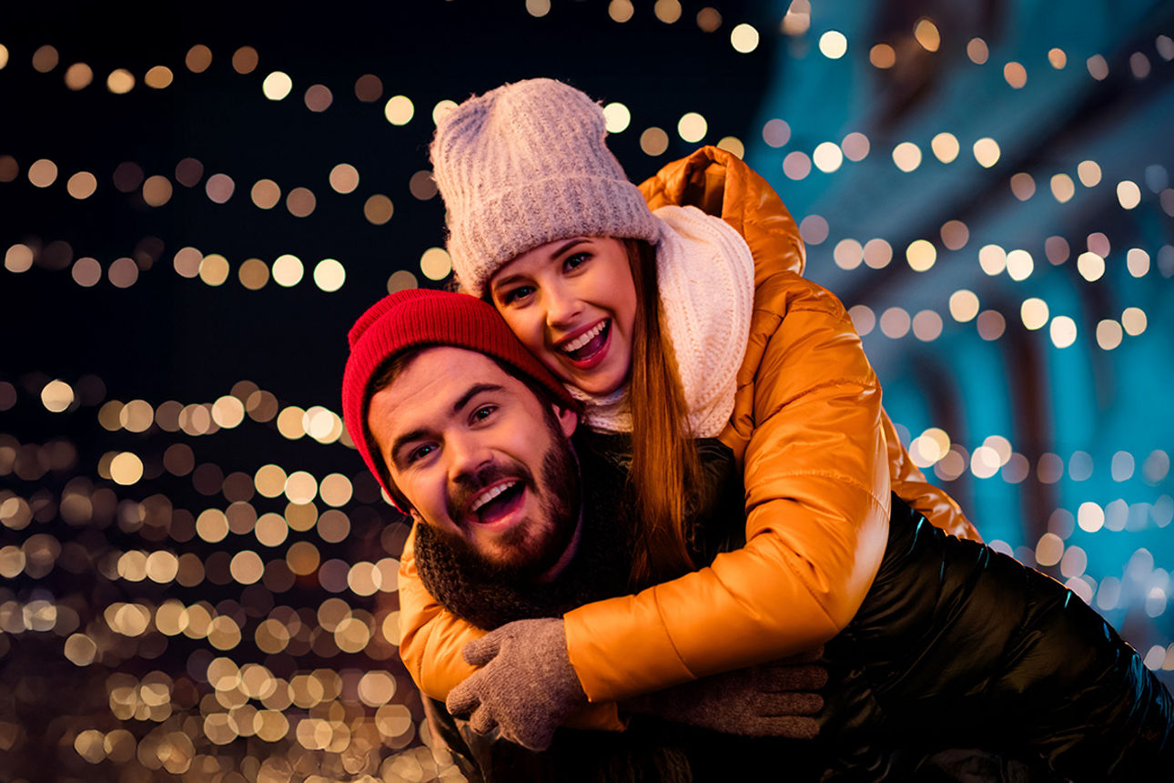Ein Mann trägt eine Frau Huckpack, hinter Ihnen leuchten viele Lichterketten