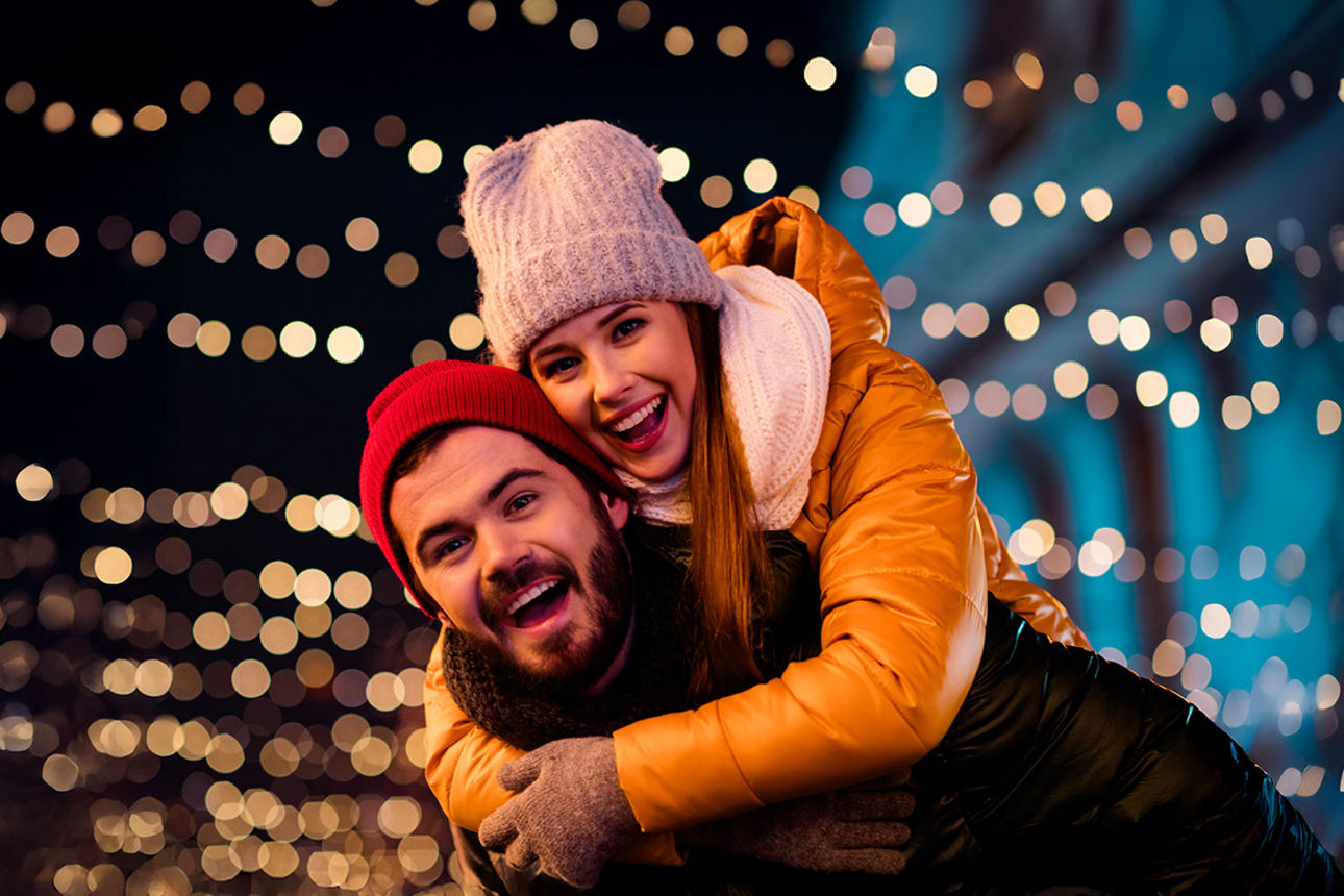 Junges Paar im Freien freut sich über den Jahreswechsel. Im Hintergrund leuchten unscharfe warme Lichterketten vor dunklem Hi