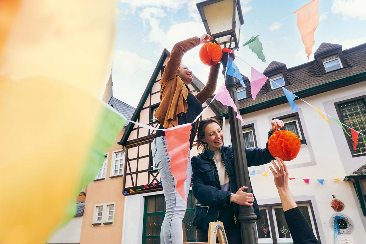 Mehrere Personen befestigen bunte Dekoration und Papierlaternen an einer Straßenlaterne in einer Straße mit Fachwerkhäusern.