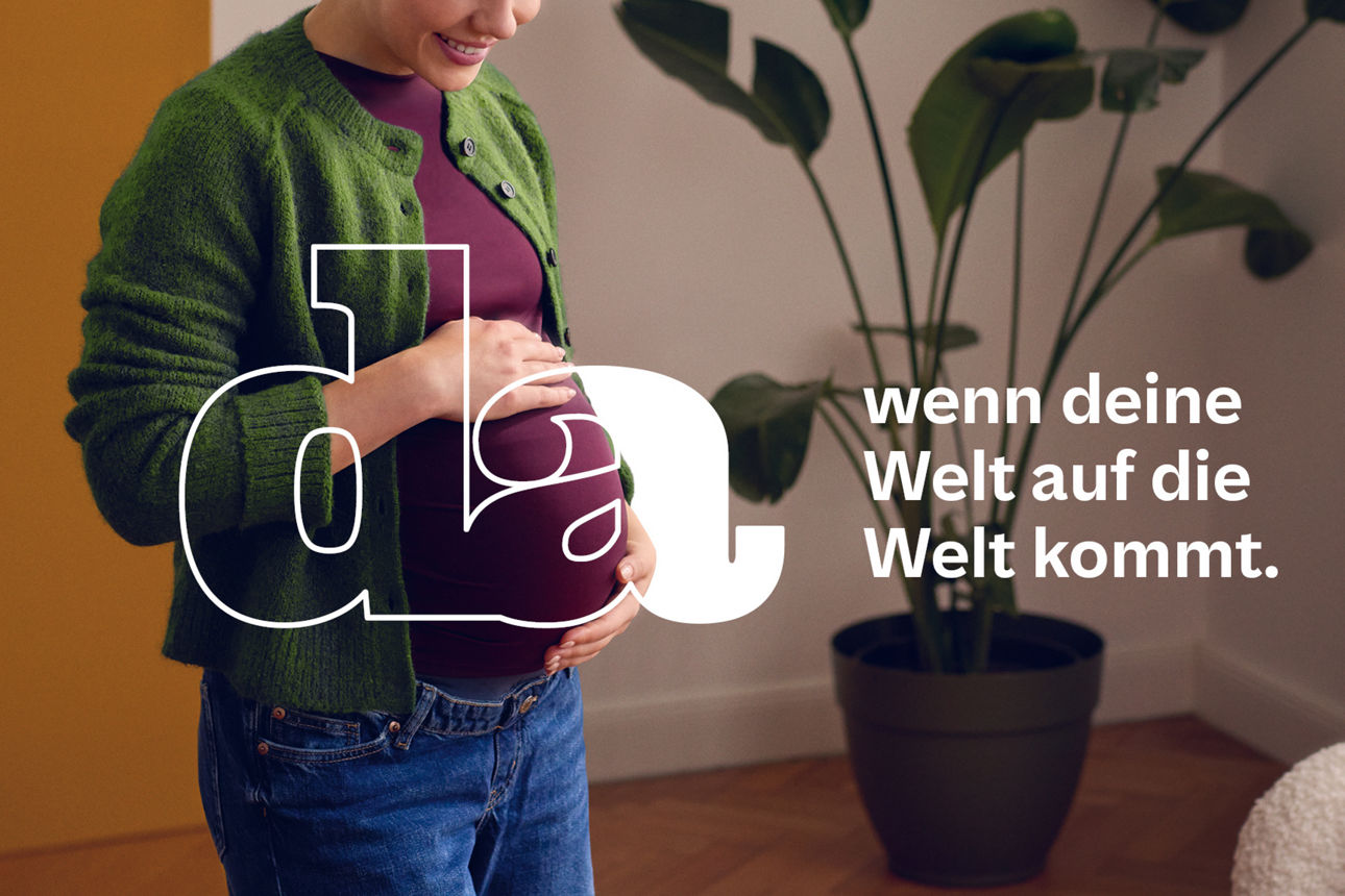 Schwangere Frau in grünem Cardigan hält ihren Bauch in einem gemütlichen Wohnzimmer mit Pflanze.