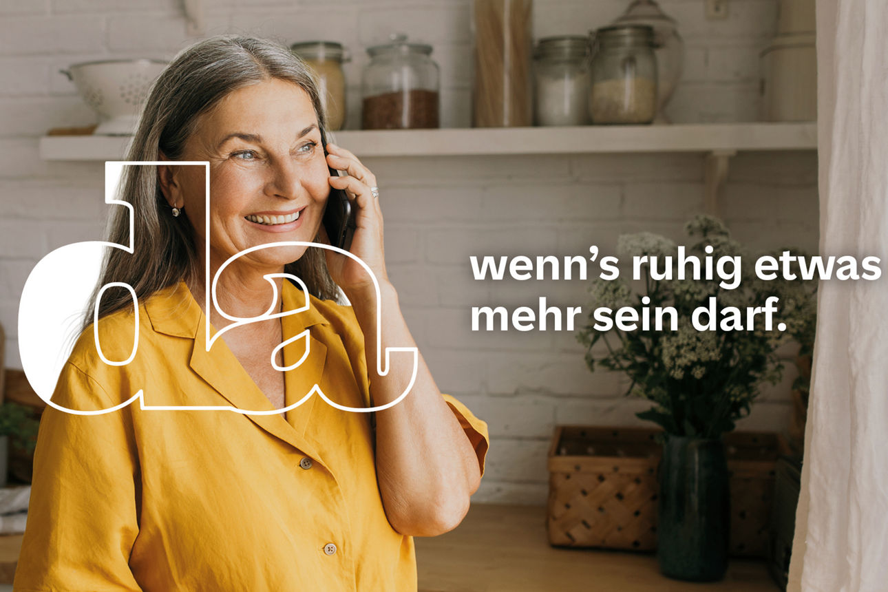 Ältere Frau mit grauem Haar und gelbem Hemd steht lächelnd in heller Küche telefoniert und hält eine Tasse in der Hand