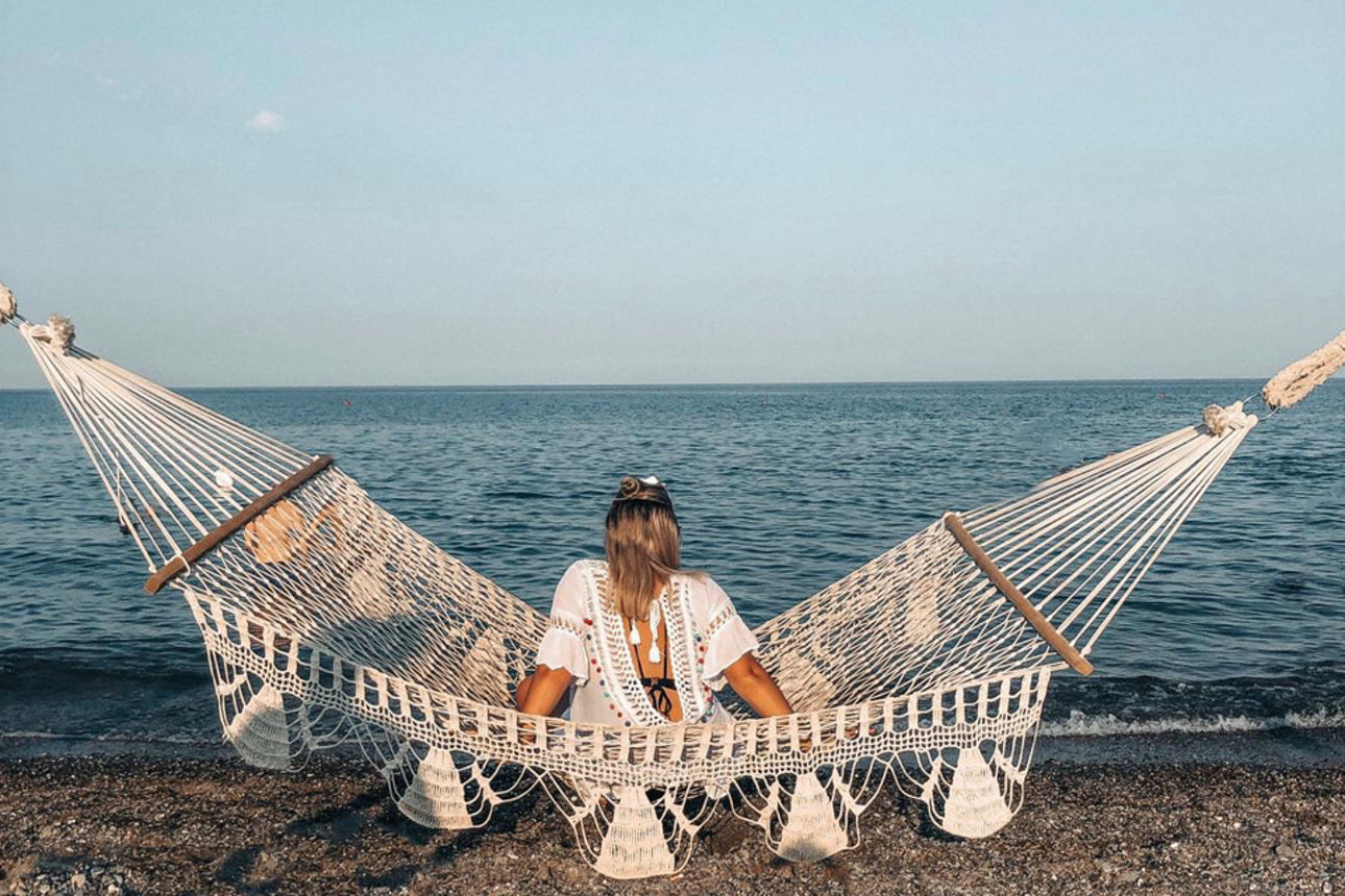 Frau sitzt in einer Hängematte am Strand und schaut aufs Meer
