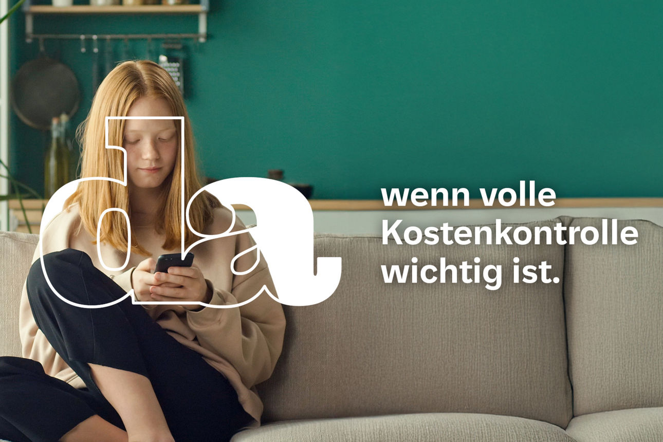 Junge Frau mit roten Haaren sitzt entspannt im Schneidersitz auf einem Sofa und schaut konzentriert auf ihr Smartphone