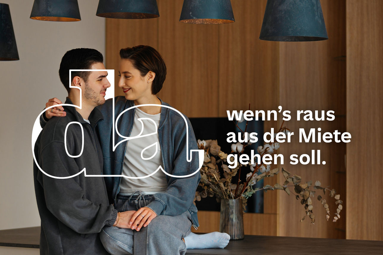 Zwei Personen sitzen eng nebeneinander auf einer Tischplatte vor einer Holzwand mit schwarzen Hängeleuchten