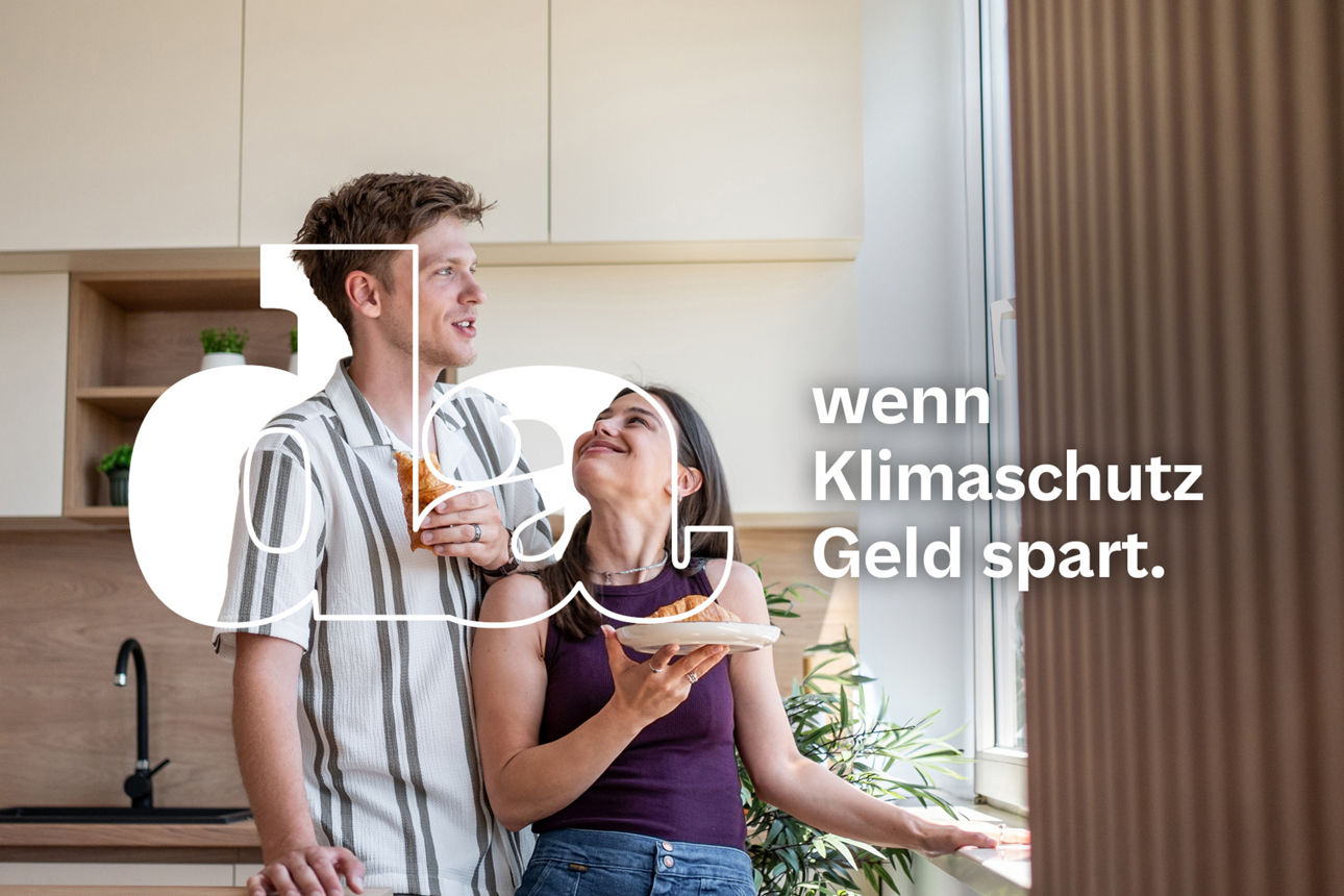 Junges Paar steht in moderner Küche und schaut sich lächelnd an sie hält ein Stück Pizza er ein Glas Saft während Sonnenlicht