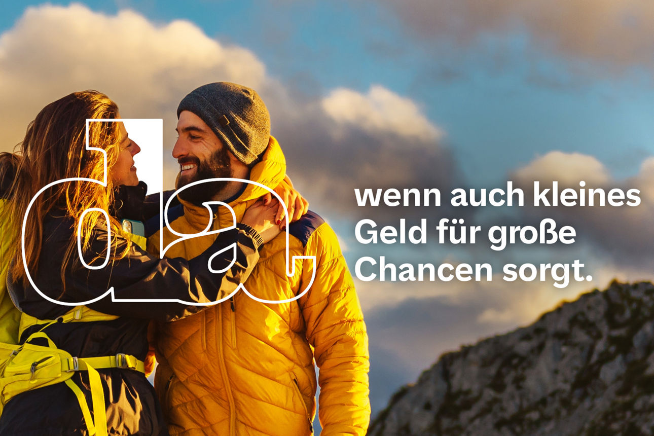 Pärchen in gelber Outdoor-Kleidung steht umarmt auf einem Berggipfel und blickt sich bei Sonnenuntergang glücklich an