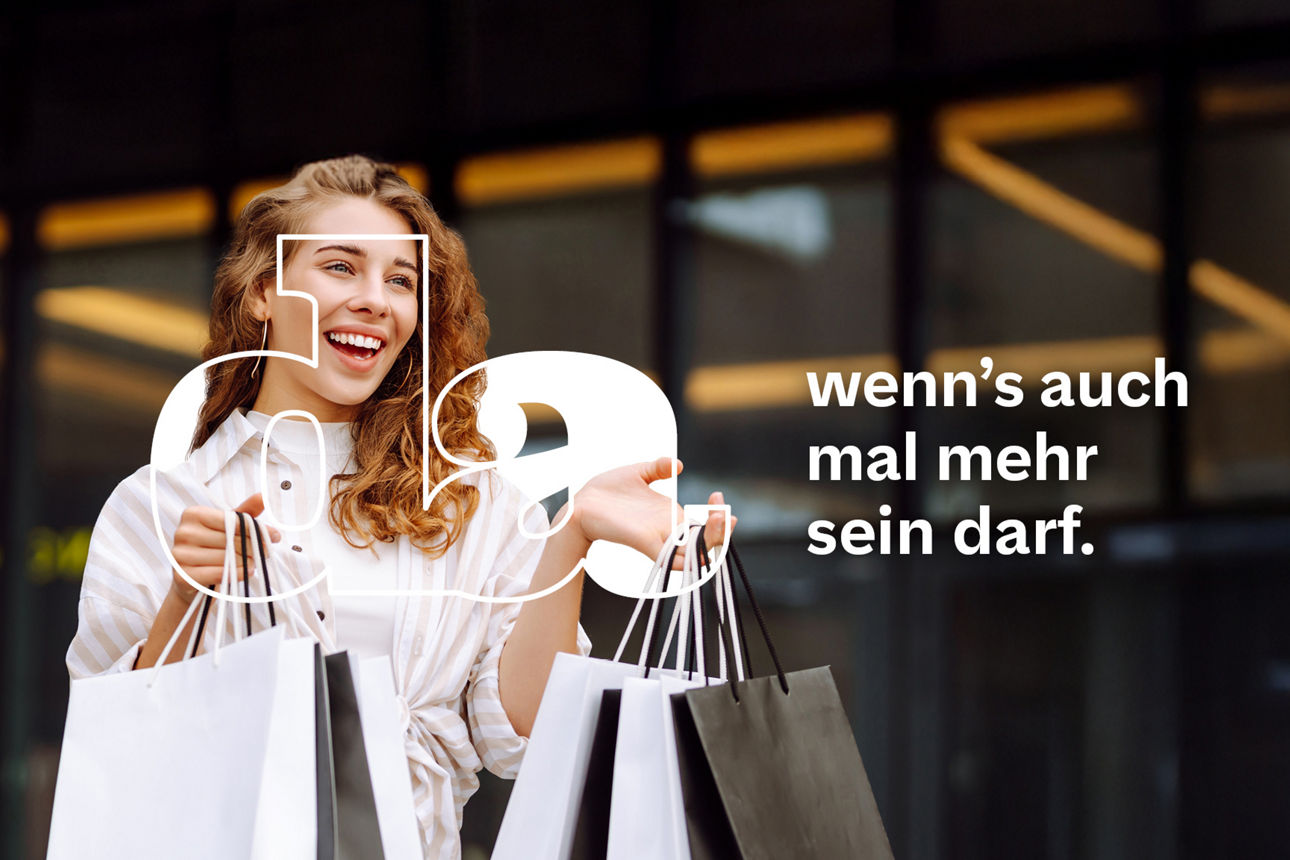 Fröhliche junge Frau mit lockigem Haar steht vor einem modernen Gebäude und hält mehrere elegante Shoppingtüten in den Händen