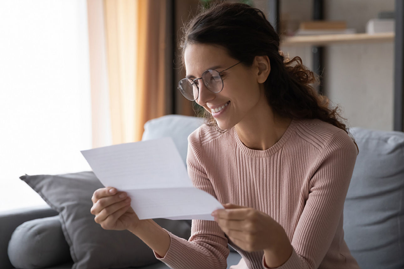 Junge Frau mit Brille liest einen Brief auf ihrem grauen Sofa
