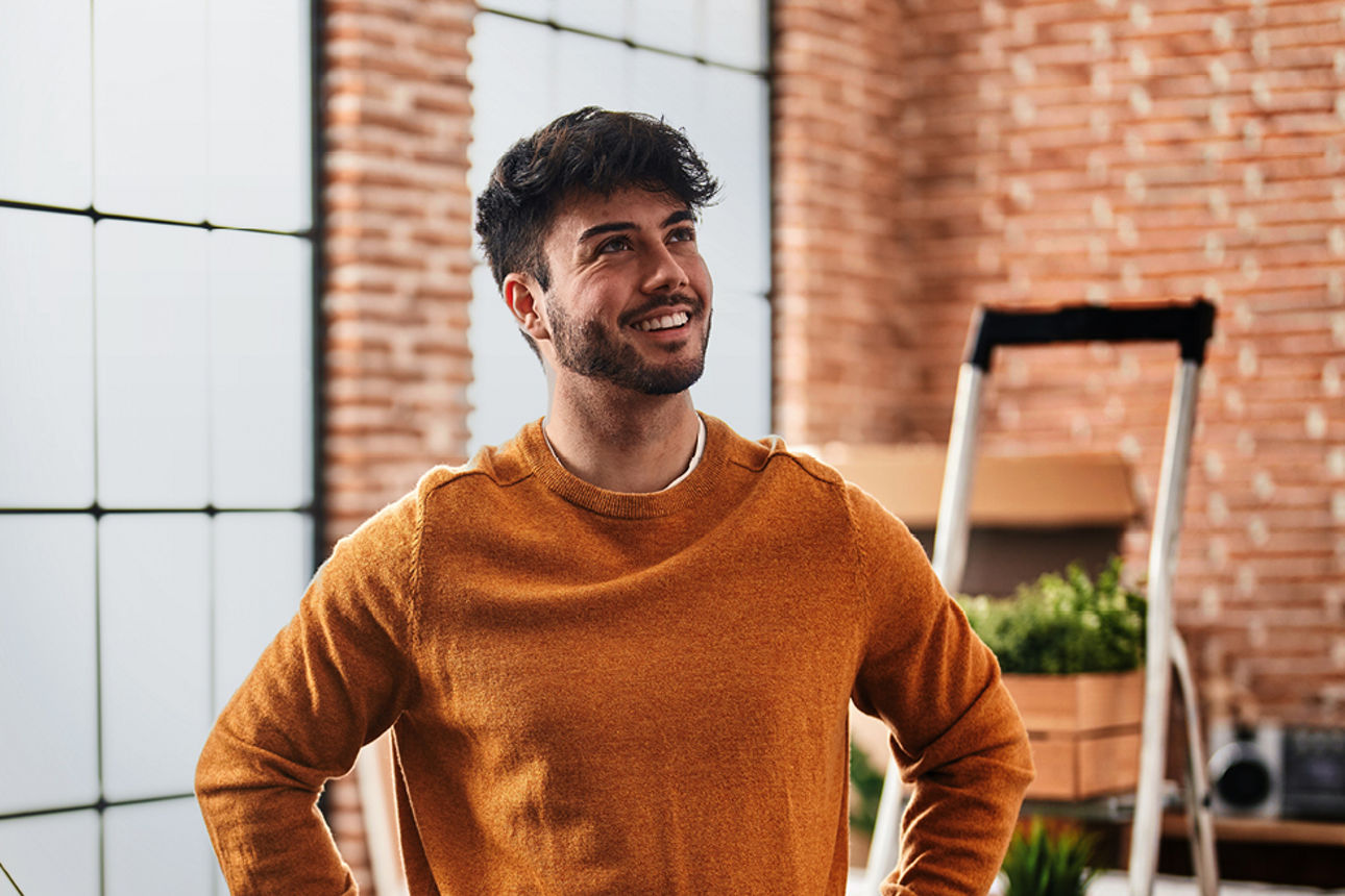Mann mit orangem Pullover steht vor einer Backsteinwand mit großen Fenstern