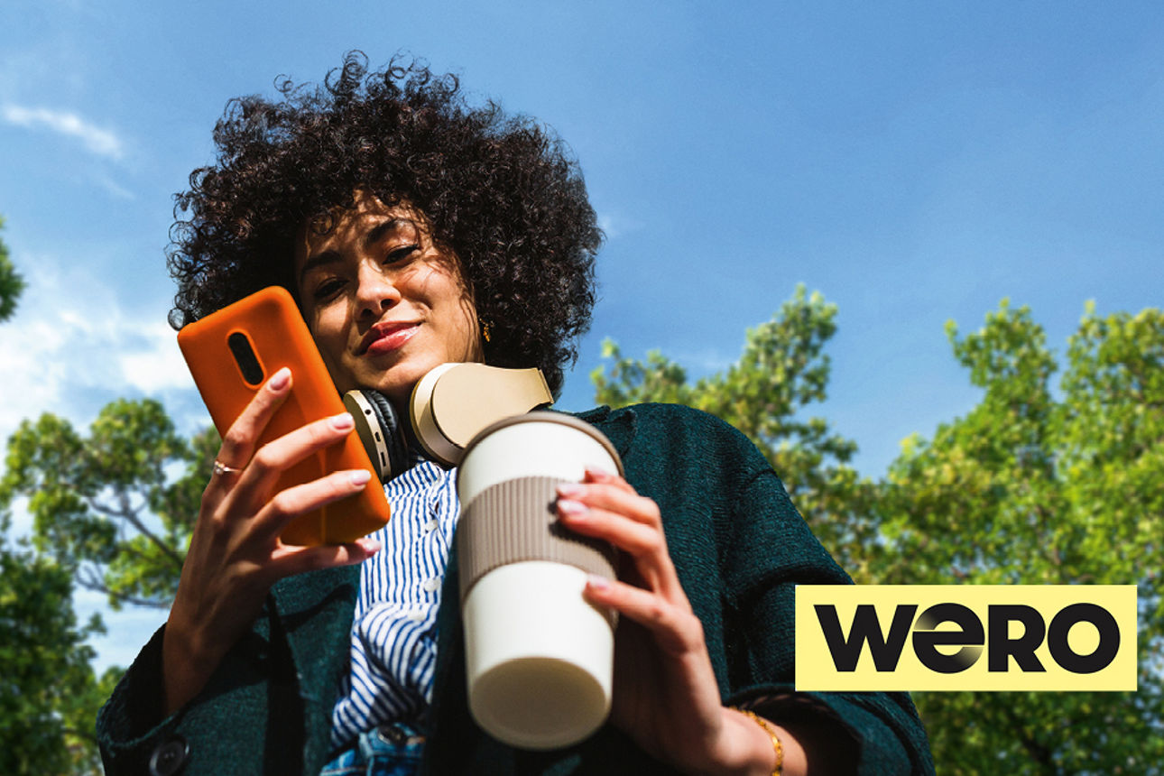 Junge Frau mit Kaffeebecher und orangenem Handy im Park. Das Wero Logo ist unten rechts zu sehen.