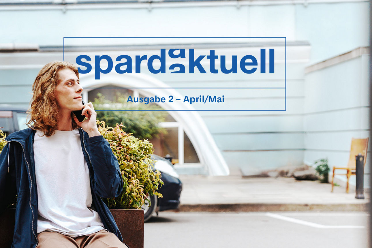 Junger Mann sitzt auf einer Bank vor einem grünen Busch und telefoniert.