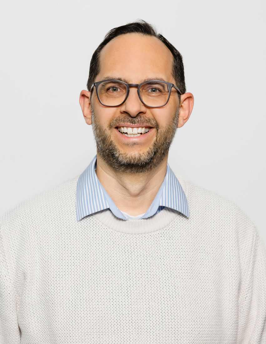 Foto unseres Vermögensberaters Ralf Kublick vor einem grauen Hintergrund in einem gestreiften Hemd und weißem Pullover