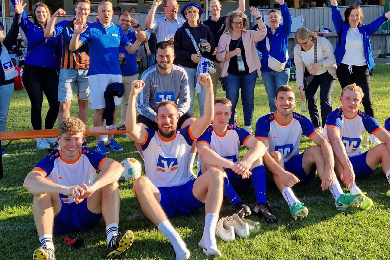 ußballteam sitzt jubelnd vor Fans auf dem Rasen; Vereinsheim mit großen Fenstern im Hintergrund, sonniger Tag.