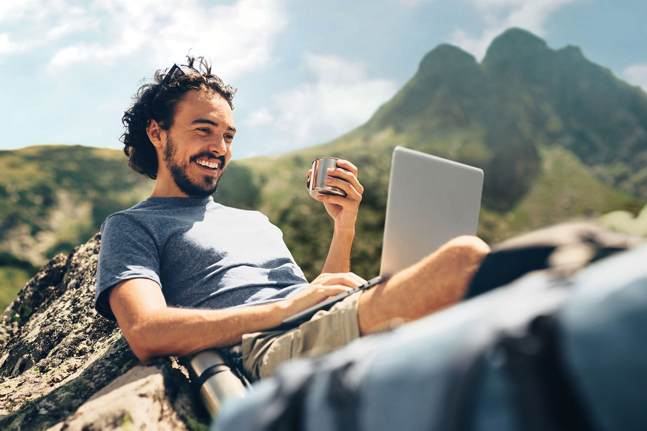 Mann sitzt an einen Stein gelehnt mit Laptop auf den Knien und Kaffeebecher in der Hand.