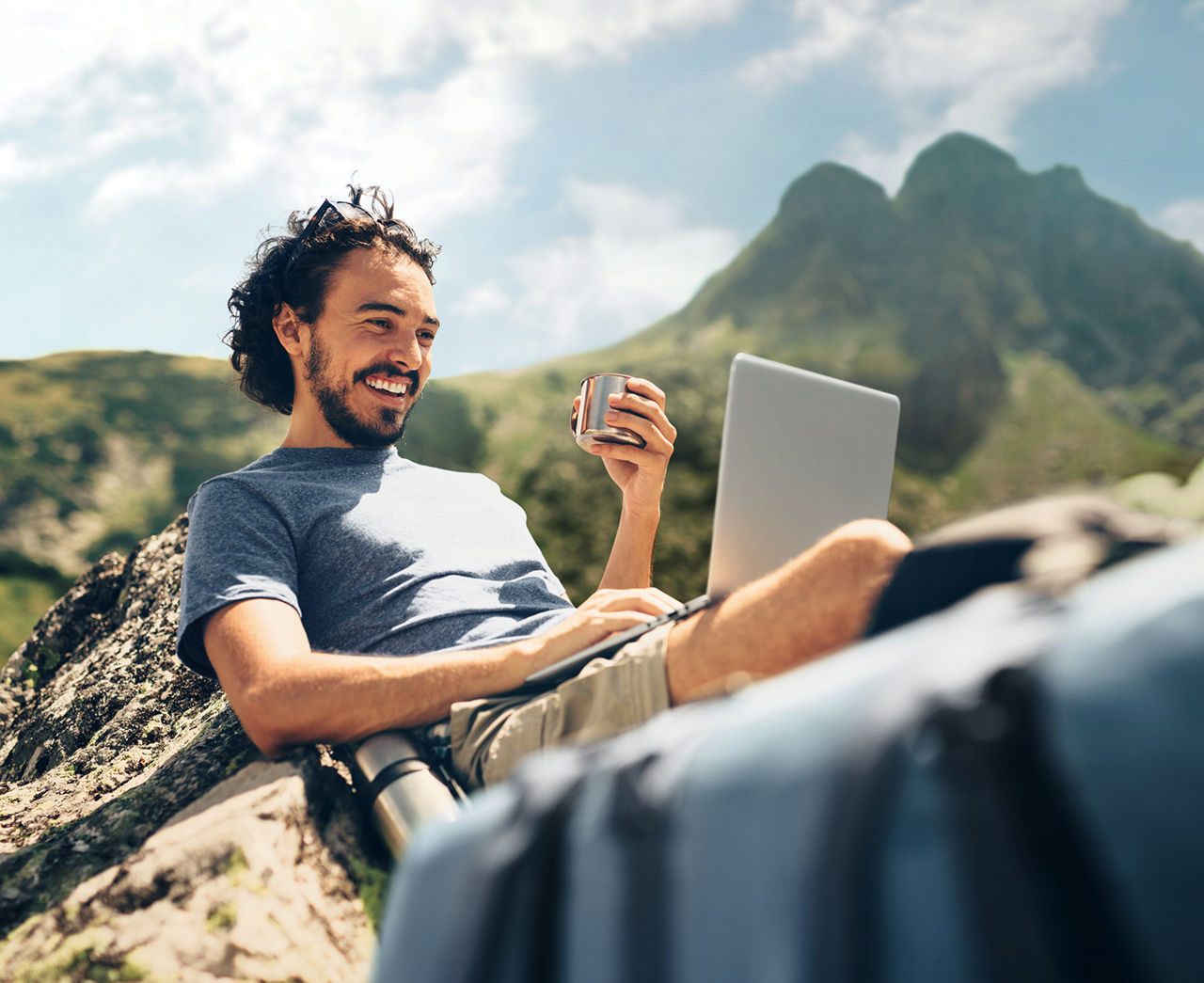 Ein junger Mann in der Natur, hinter ihm eine Landschaft mit Bergen, dabei hat er ein Laptop auf seinem Schoß. 