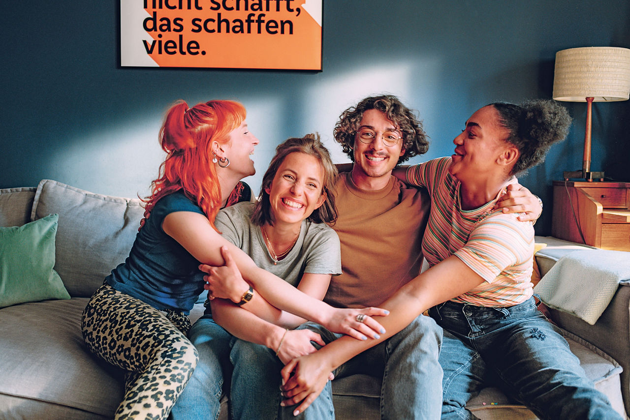 Vier junge Menschen sitzen auf einer Couch, nehmen sich in den Arm und lächeln.